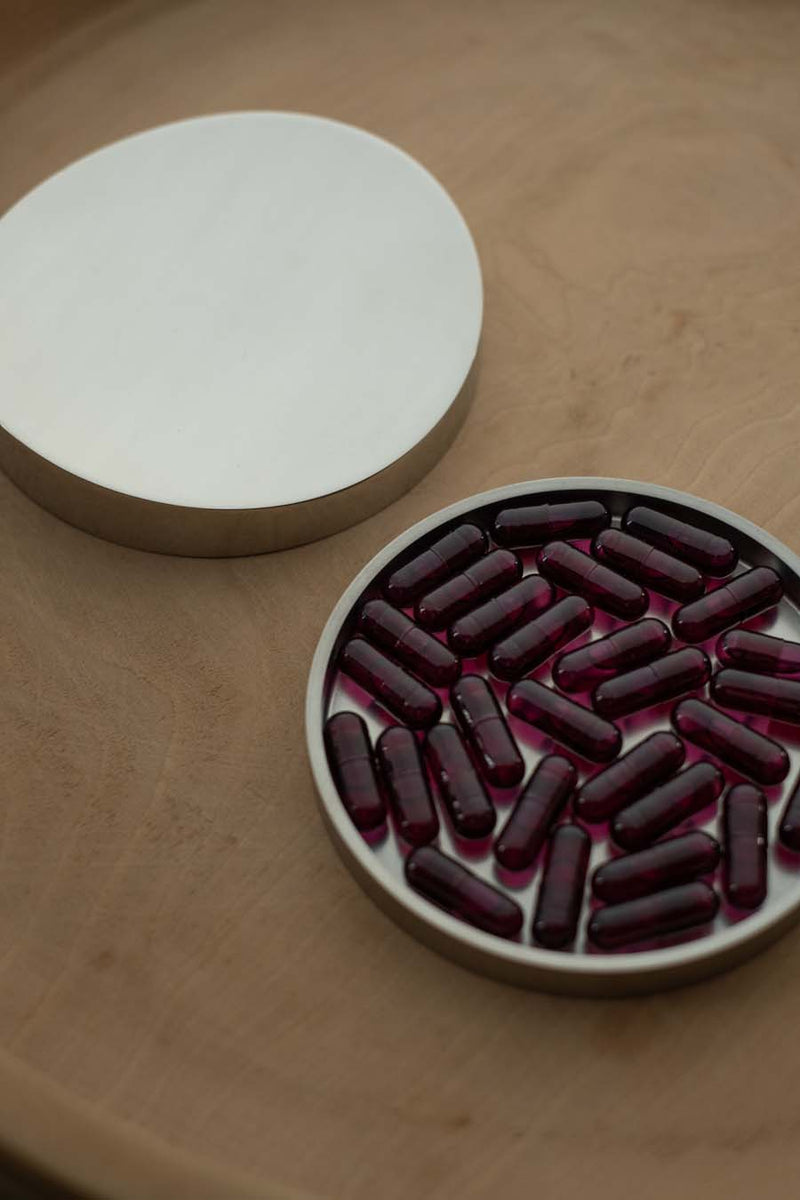 Silver vitamin storage container on a wooden surface with magenta food supplement capsules inside