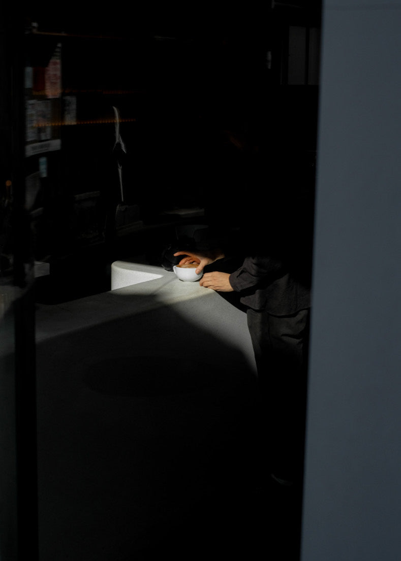 Person sitting at a table in a dimly lit room with a coffee cup on the table.