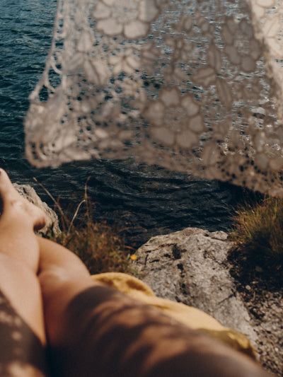 Person sitting on a rock by the ocean with a lace shade curtain in the foreground