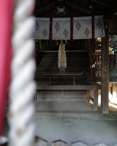 Traditional wooden shrine with white curtains and a tree inside, viewed through a red and white torii gate.
